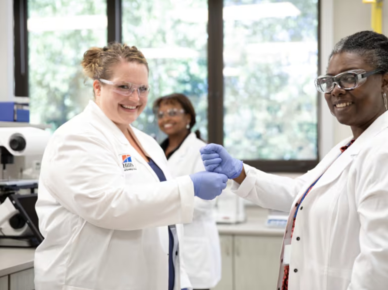 three smiling women, scientists in white coats and protective glasses
