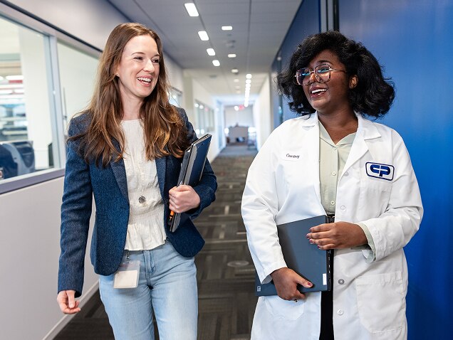 Two women, one in casual clothes and one in a white lab coat, walk and talk in a Colgate-Palmolive building corridor, smiling and holding notebooks.