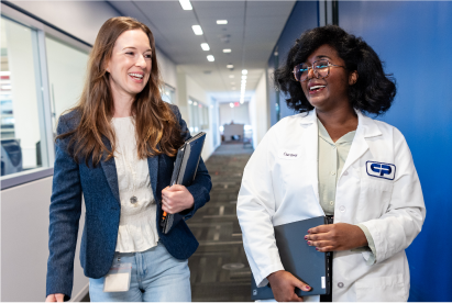 Two women, one in casual clothes and one in a white lab coat, walk and talk in a Colgate-Palmolive building corridor, smiling and holding notebooks.