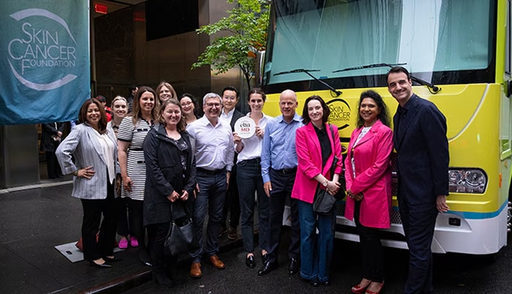 Group of people smiling at the camera while standing in front of a yellow vehicle as part of an event for "Skin Cancer Foundation".