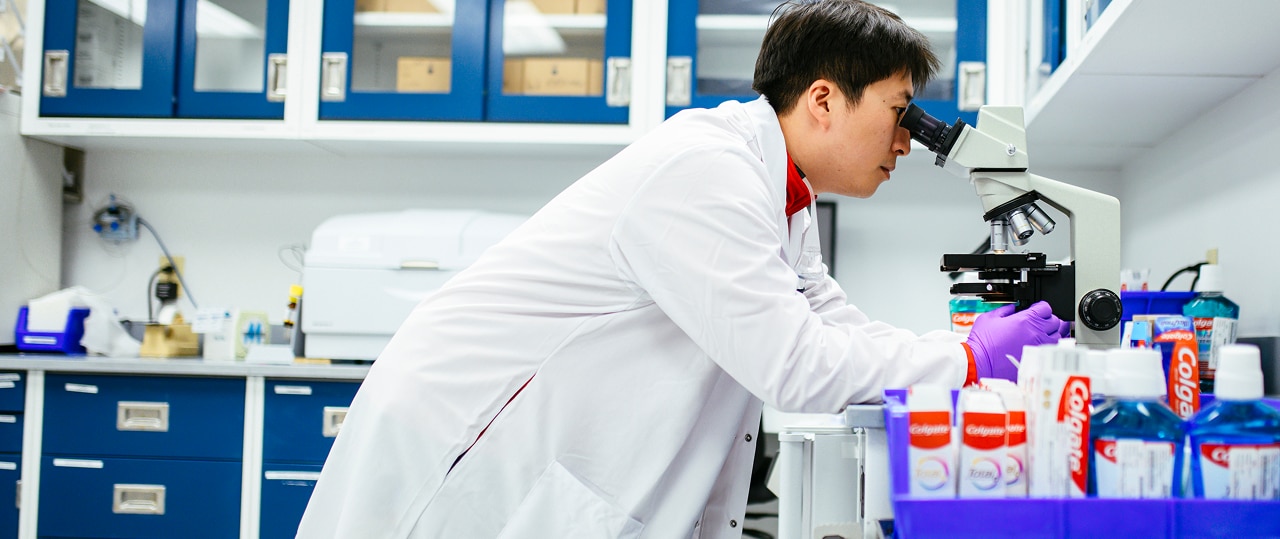 Man looking into a microscope while in a laboratory alongside a group of Colgate products.