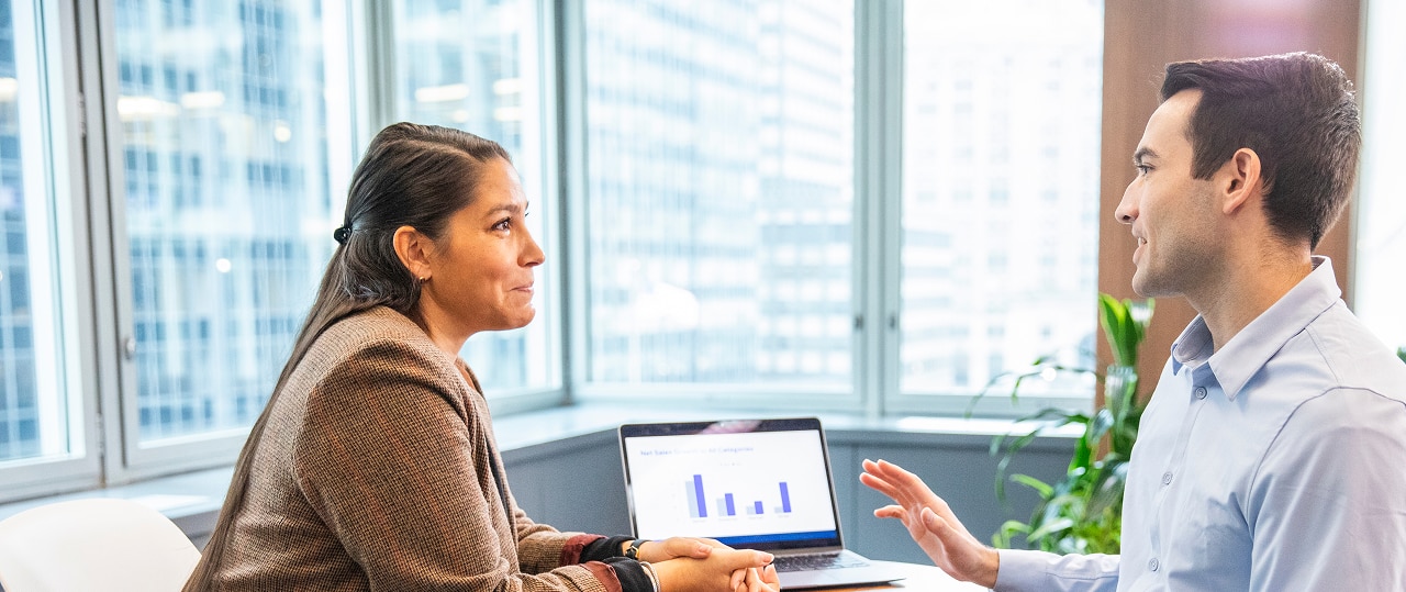 Woman listening to man explain digital data findings in an office setting next to a laptop.