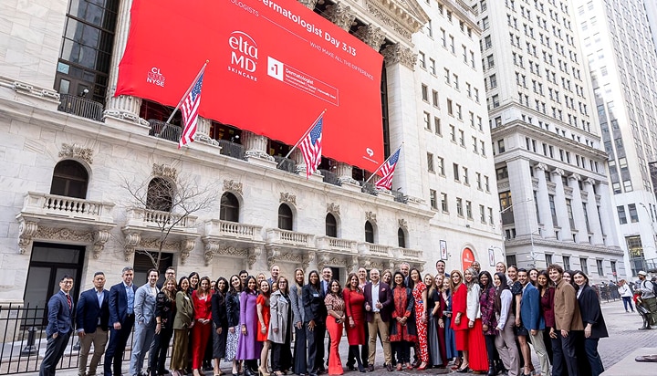 Large group of people gathered outside of a building as part of a celebration for National Dermatologist Day. A bright red banner showcases the logo of EltaMD, who created the day, alongside United States flags.