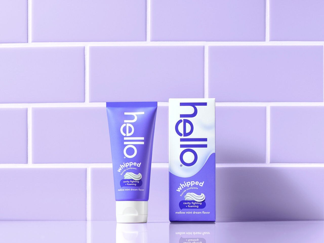 Purple and white hello Whipped toothpaste tube and box in front of purple tiled wall.