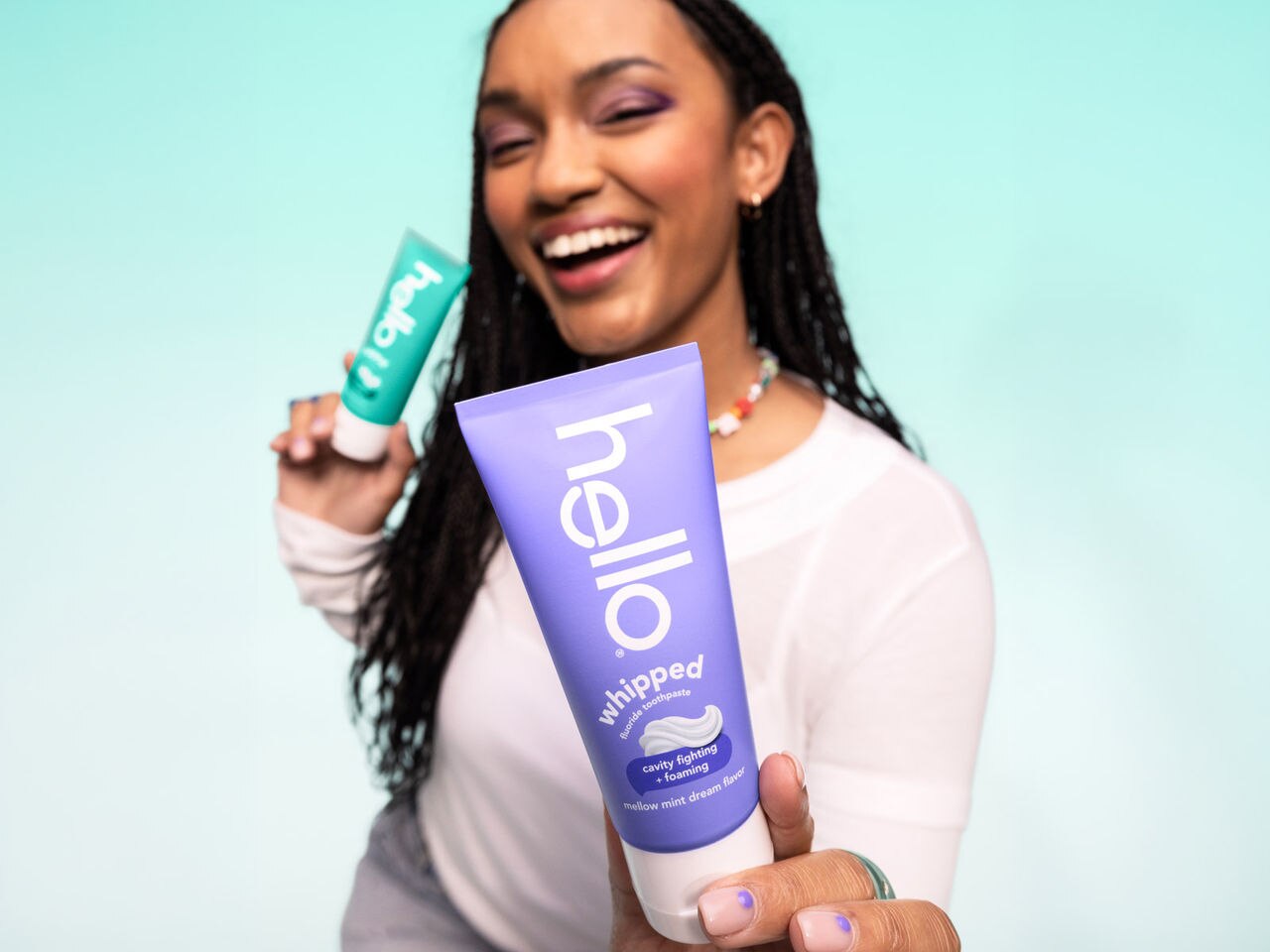 Smiling woman with two hello toothpaste tubes holds a purple tube close to the viewer.