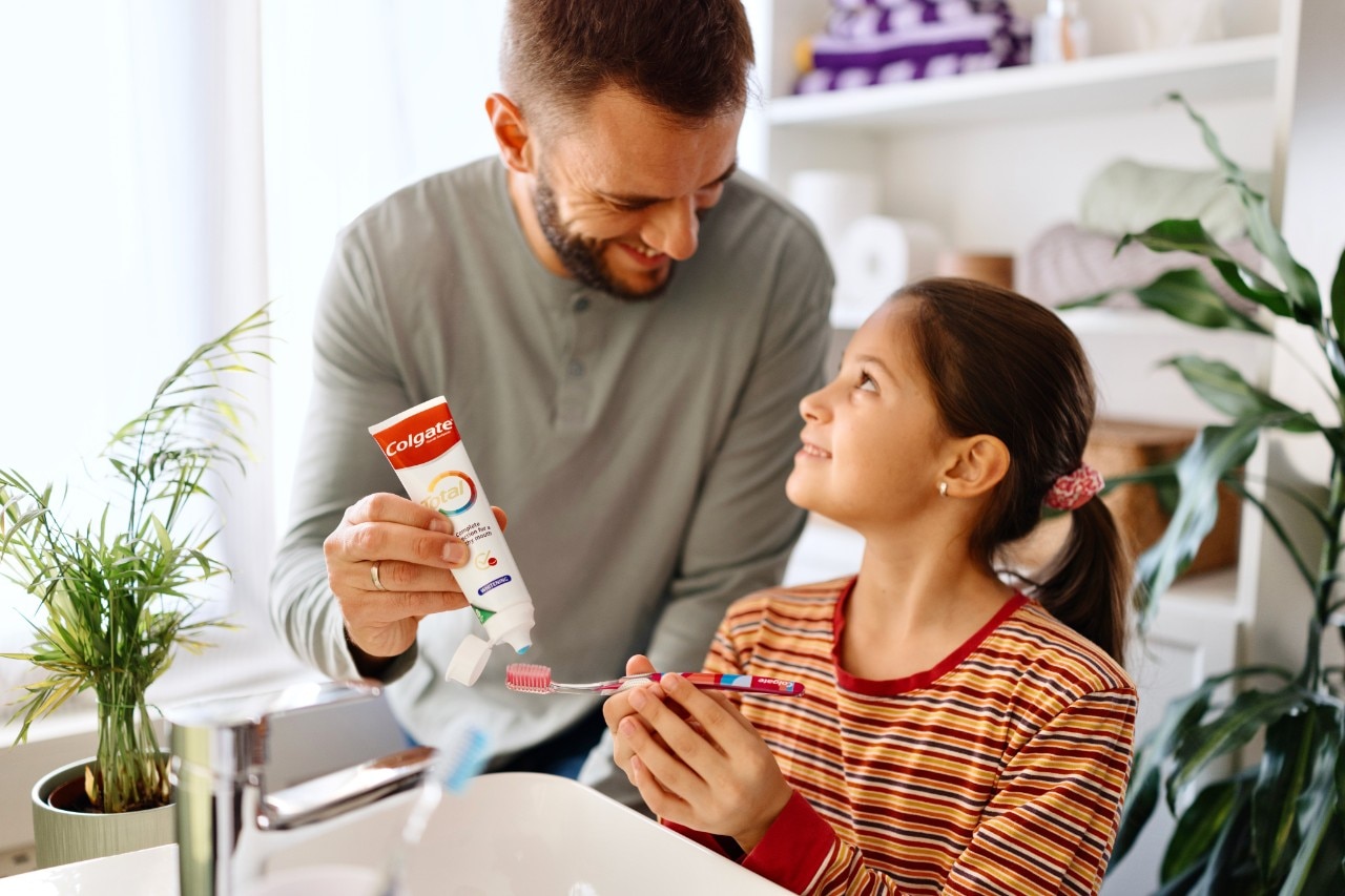 A man applying toothpaste to a girl's toothbrush at a sink, with plants in the background.