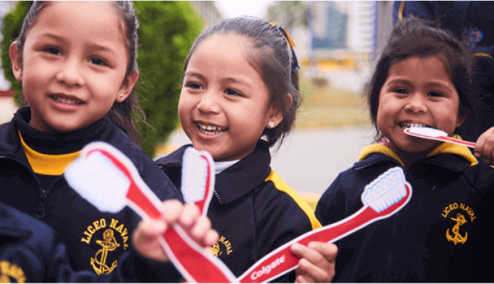 Three smiling young girls wearing school uniforms holding Colgate toothbrushes outdoors.