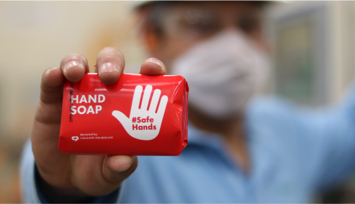 Person wearing a mask holds a red bar of hand soap labeled "HAND SOAP #Safe Hands" for cleaning and hygiene.